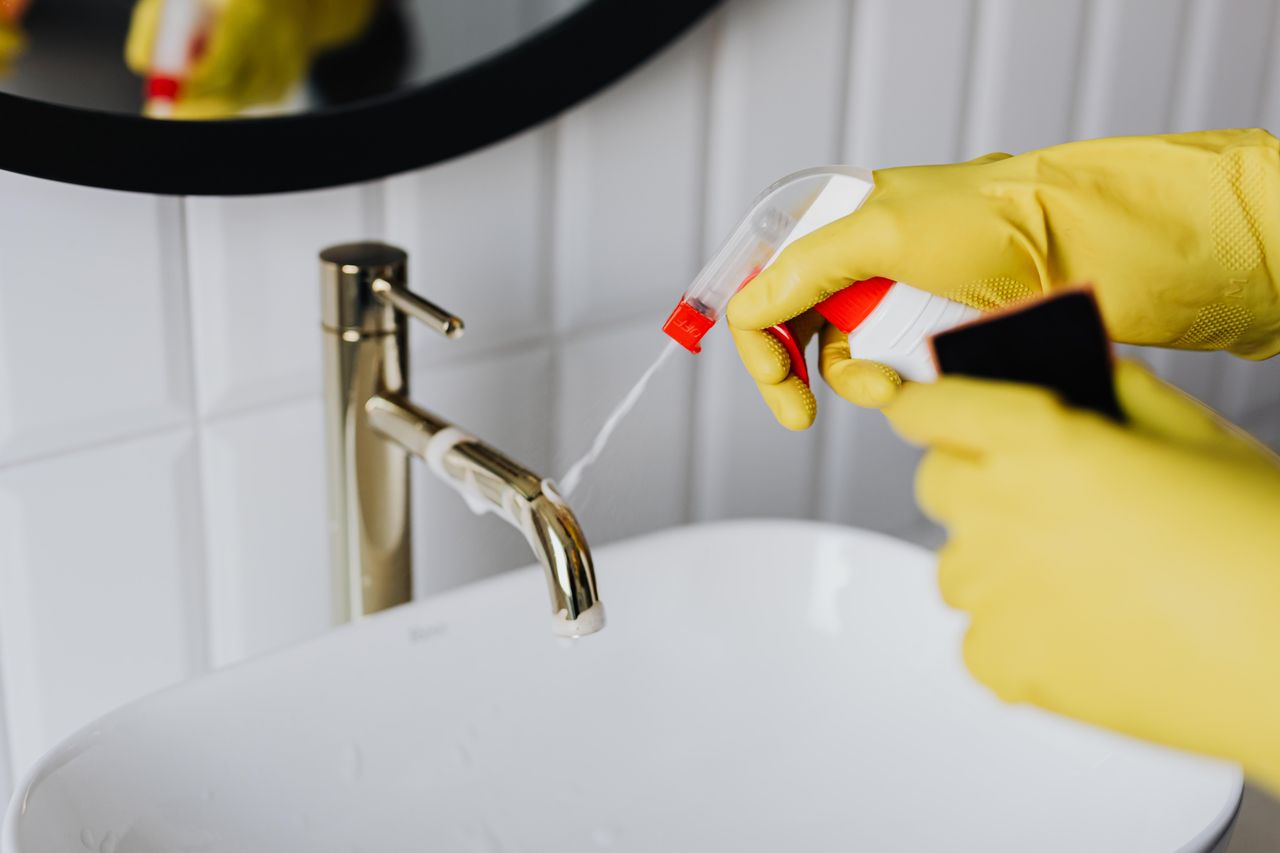 services-04 From above anonymous housekeeper in yellow rubber gloves spraying cleaner to chrome water faucet in bathroom