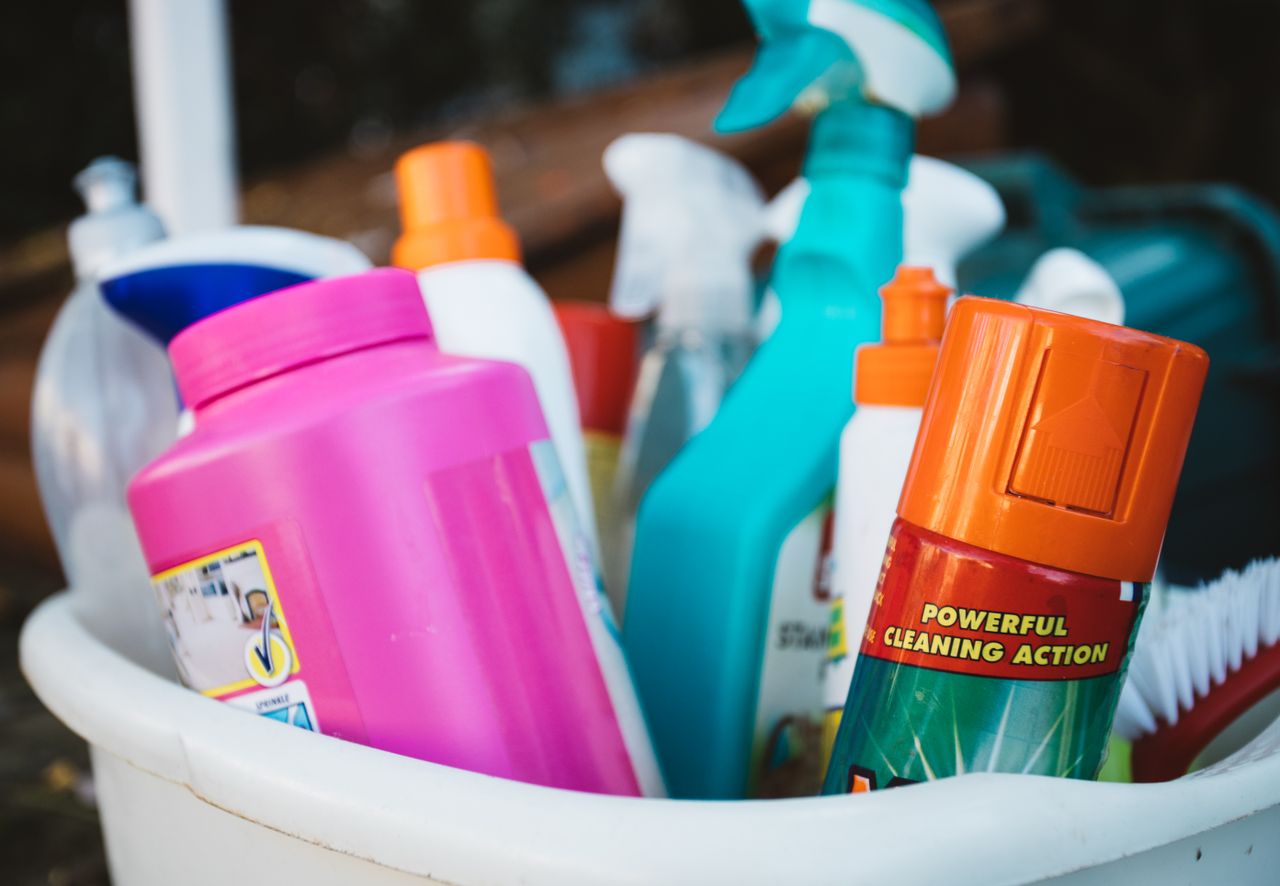 Gros plan sur divers produits de nettoyage dans un seau à l’extérieur, mettant en avant différents détergents et sprays.